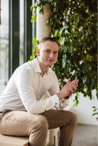 Tobias Lechner sitzt im Büro von Lechner IT-Dienstleistungen auf einer Bank. Er trägt ein helles Hemd und beige Hosen und blickt zur Kamera.